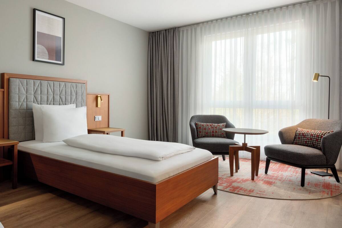Een hotelkamer met een bed, twee fauteuils, een tafeltje, een lamp en een raam met gordijnen.