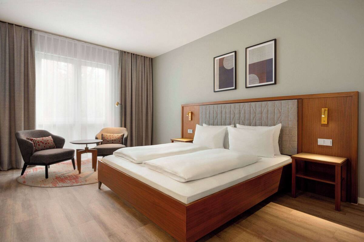 Een hotelkamer met een groot bed, twee nachtkastjes, een stoel, een klein tafeltje en een groot raam met gordijnen.