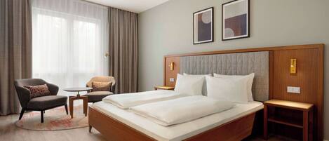 A hotel room with a large bed, two bedside tables, a chair, a small table, and a large window with curtains.