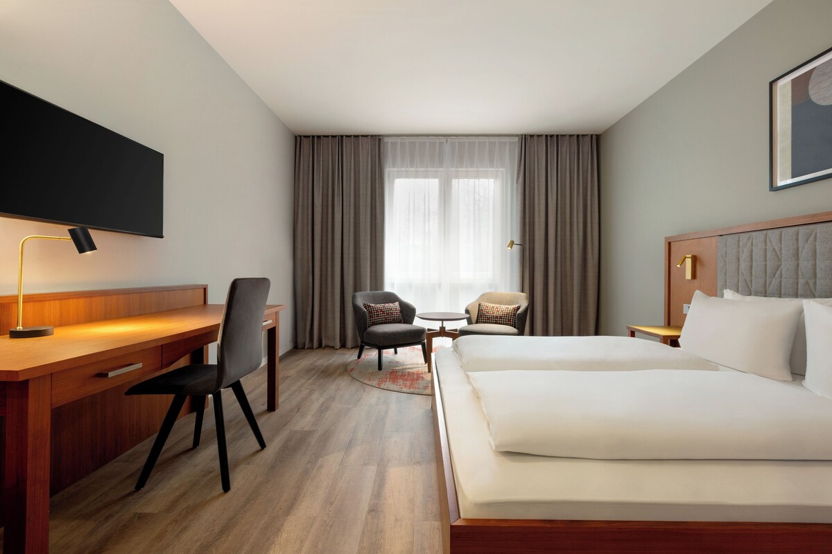 Een hotelkamer met een bed, bureau, twee stoelen, een televisie en een raam met gordijnen.