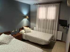 A bedroom with two beds, a bedside table, a lamp, a window with curtains, and a television. - Hostal Cal Jaume (Mollerussa)