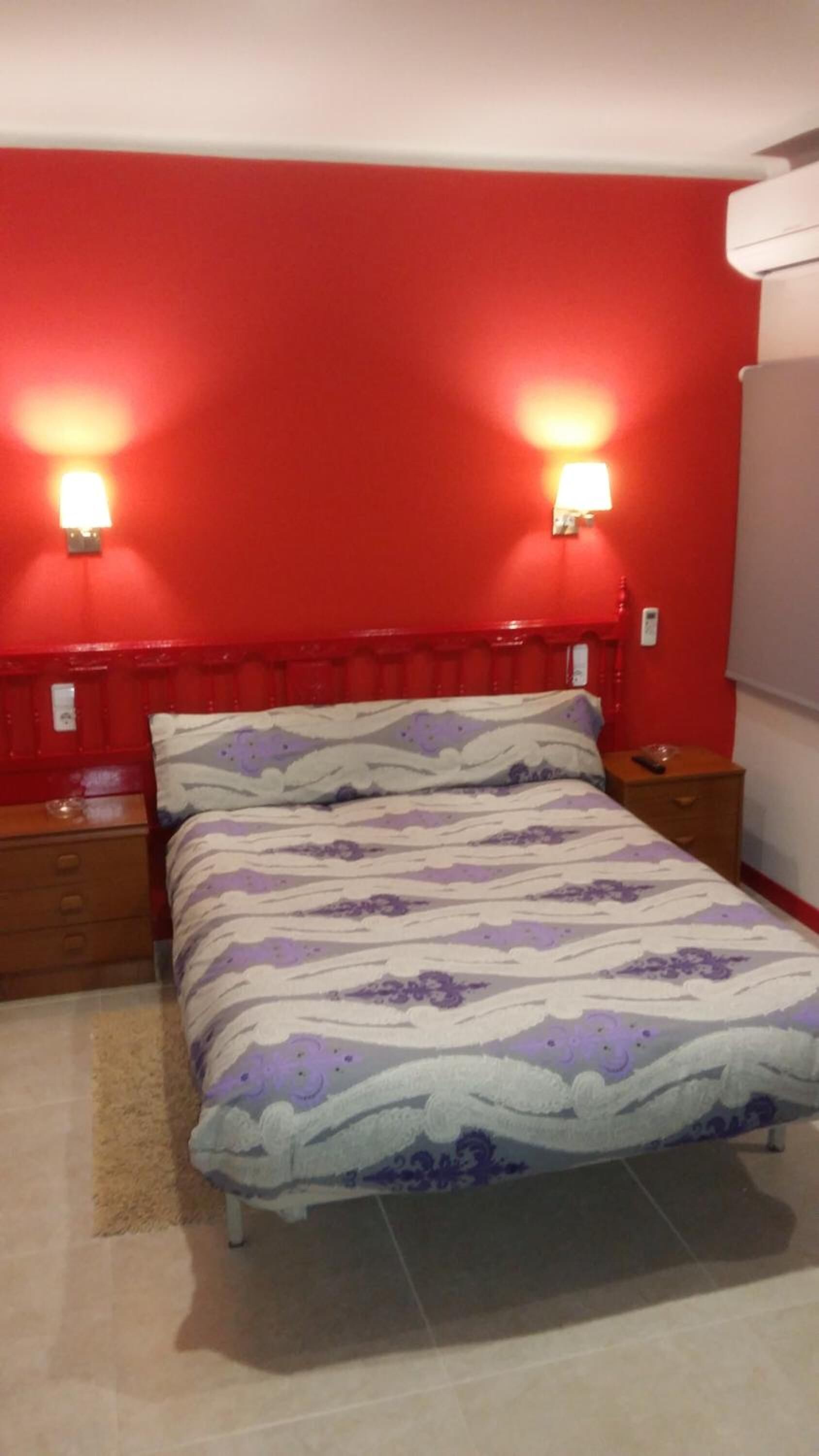Un dormitorio con cabecera roja, dos lámparas de noche, cama con colcha estampada y mesita de noche con un control remoto.