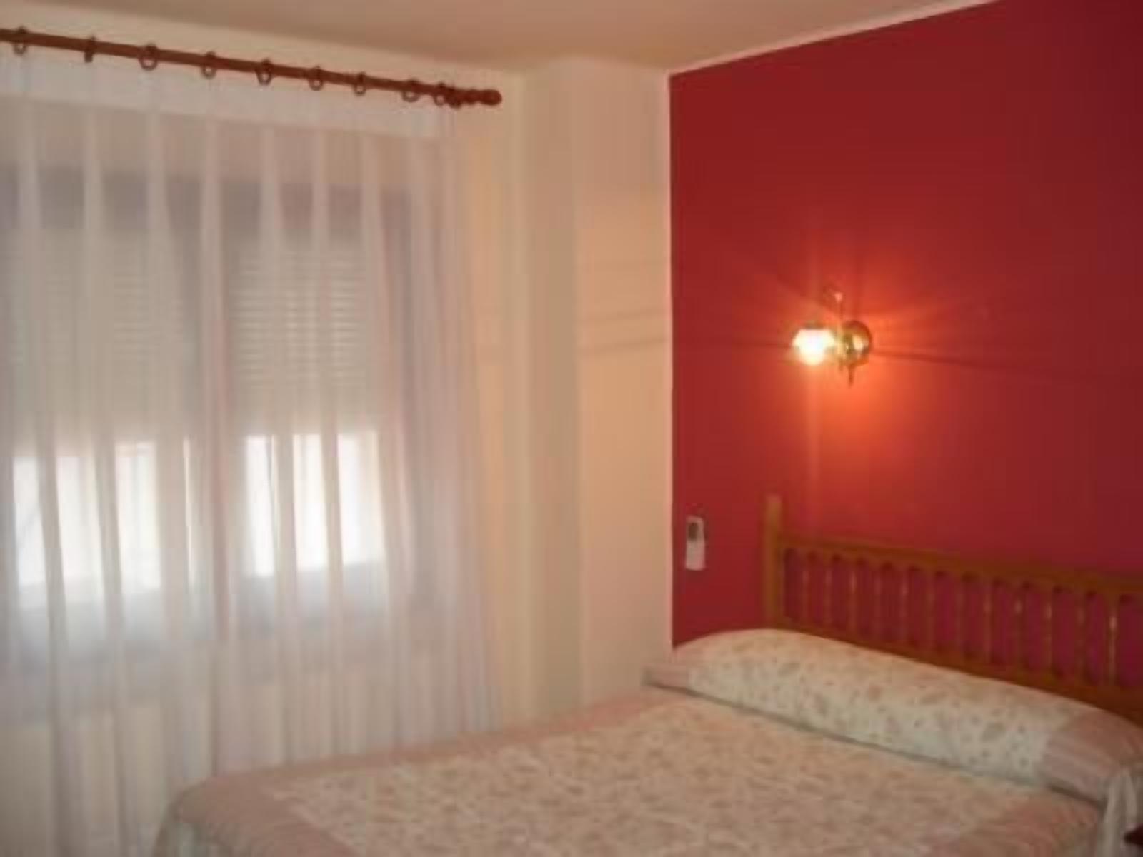 Un dormitorio con cama, ventana con cortinas de gasa y lámpara fijada a la pared.