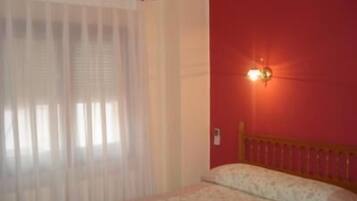 Un dormitorio con cama, ventana con cortinas de gasa y lámpara fijada a la pared.