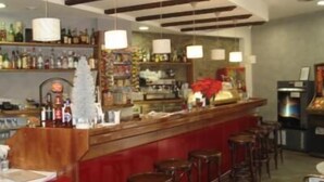 Eine Bar mit einer Holztheke, mehreren hängenden Pendelleuchten und einer Auswahl an Flaschen und Gläsern auf Regalen.