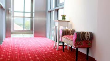 A hallway with a red carpet, a white chair with floral upholstery, and a small table with a vase of flowers.
