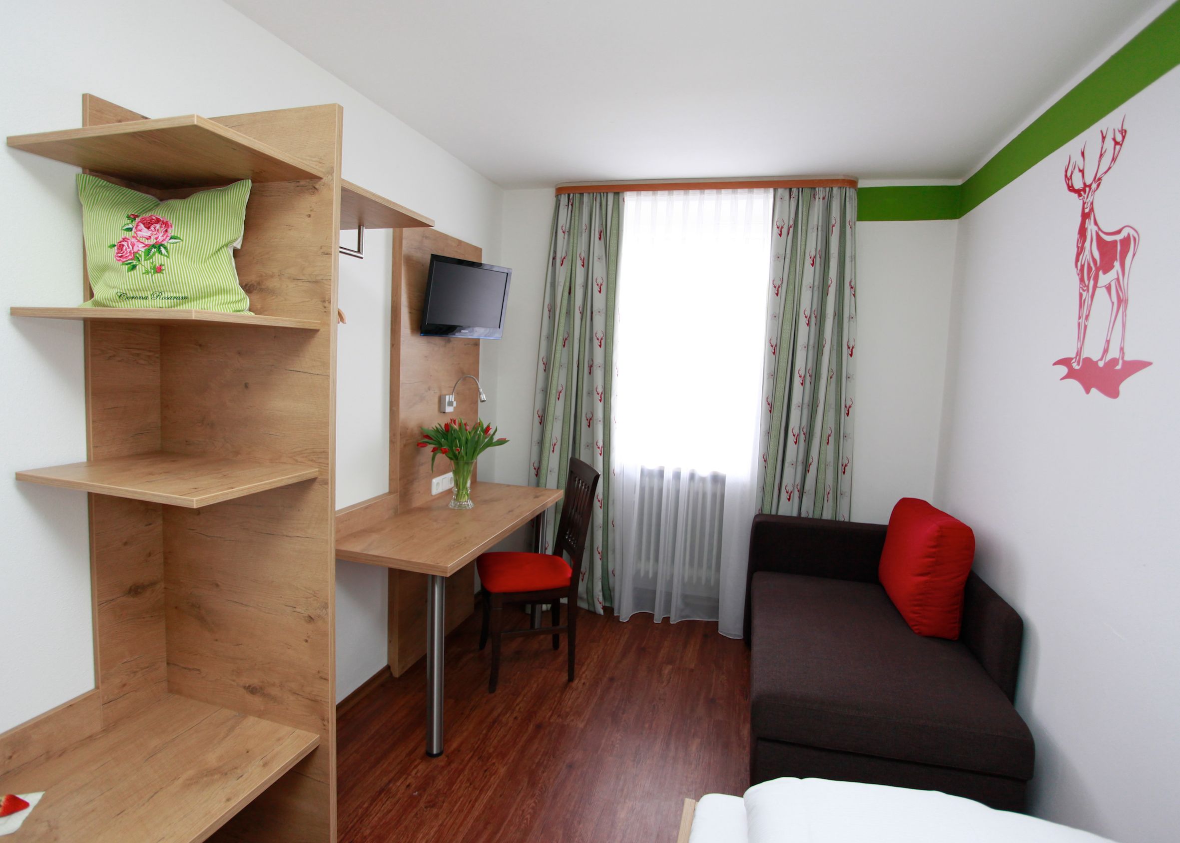Een kleine kamer met een houten bureau, een televisie, een rode stoel, een bruine bank en een bed.