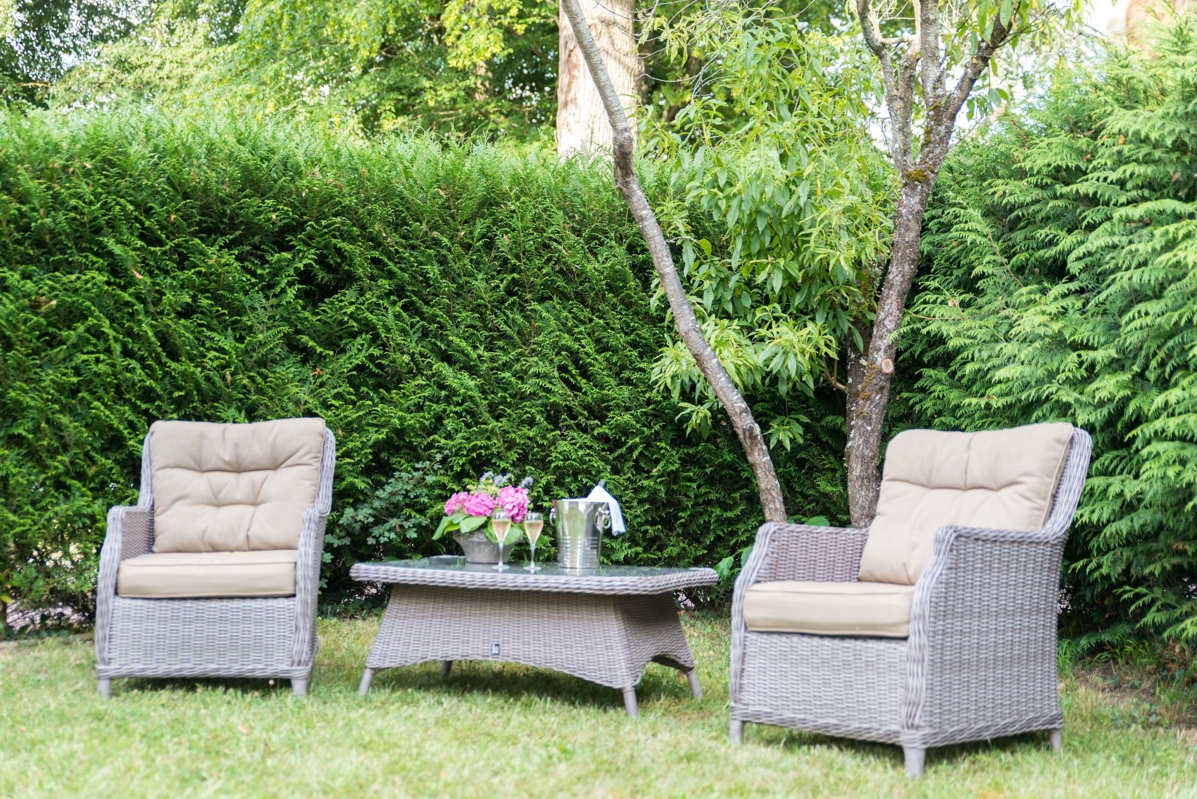 A patio with two wicker chairs and a table, surrounded by lush greenery.