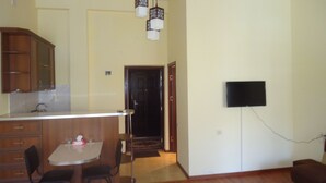 A kitchen with wooden cabinets, a white countertop, and a dining area with two chairs and a table. There is a television mounted on the wall and a door leading to another room. - Apartments Tigran Petrosyan (Yerevan)