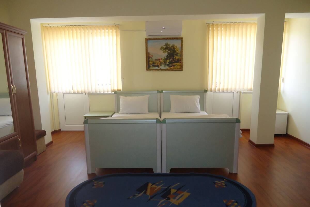 A bedroom with a double bed, two bedside tables, a small bedside table, a radiator, a painting on the wall, and a blue rug with a geometric pattern.