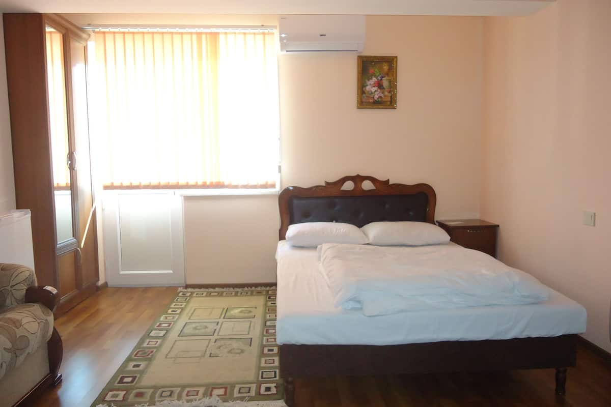 A bedroom with a bed, a nightstand, a chair, a window with curtains, and a wall-mounted air conditioner.
