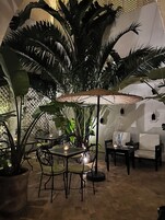 Un patio interior con mesas y sillas, rodeado de plantas grandes y un paraguas decorativo.