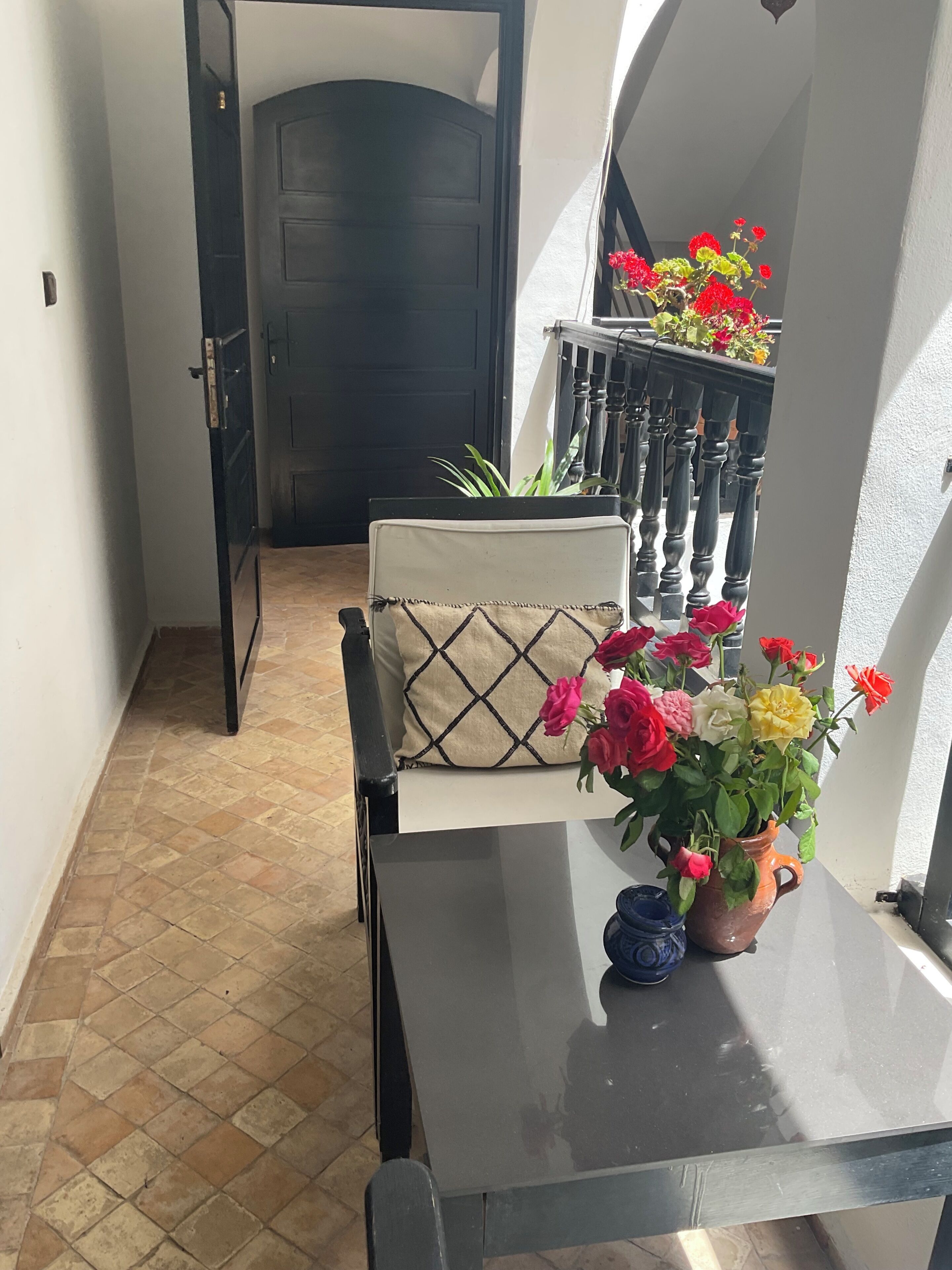 Un pasillo con una puerta, una silla con cojín, una mesa con un jarrón de flores y una escalera con barandilla.