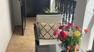 Un pasillo con una puerta, una silla con cojín, una mesa con un jarrón de flores y una escalera con barandilla.
