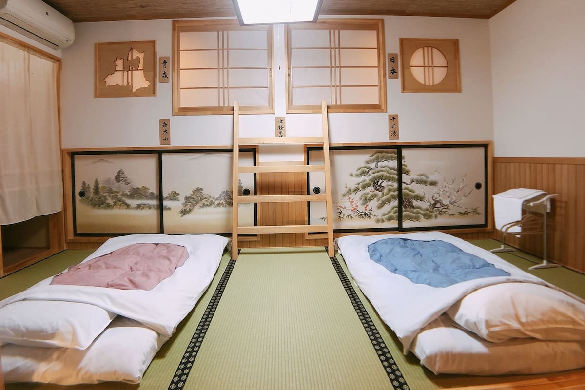 A traditional Japanese-style room with tatami flooring, two beds with shoji screens, and artwork depicting nature scenes.