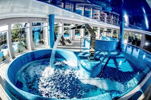 A large indoor swimming pool with a waterfall feature, surrounded by a covered area with seating and a view of the outside. - Tristan Hotel & Spa (Sztutowo)