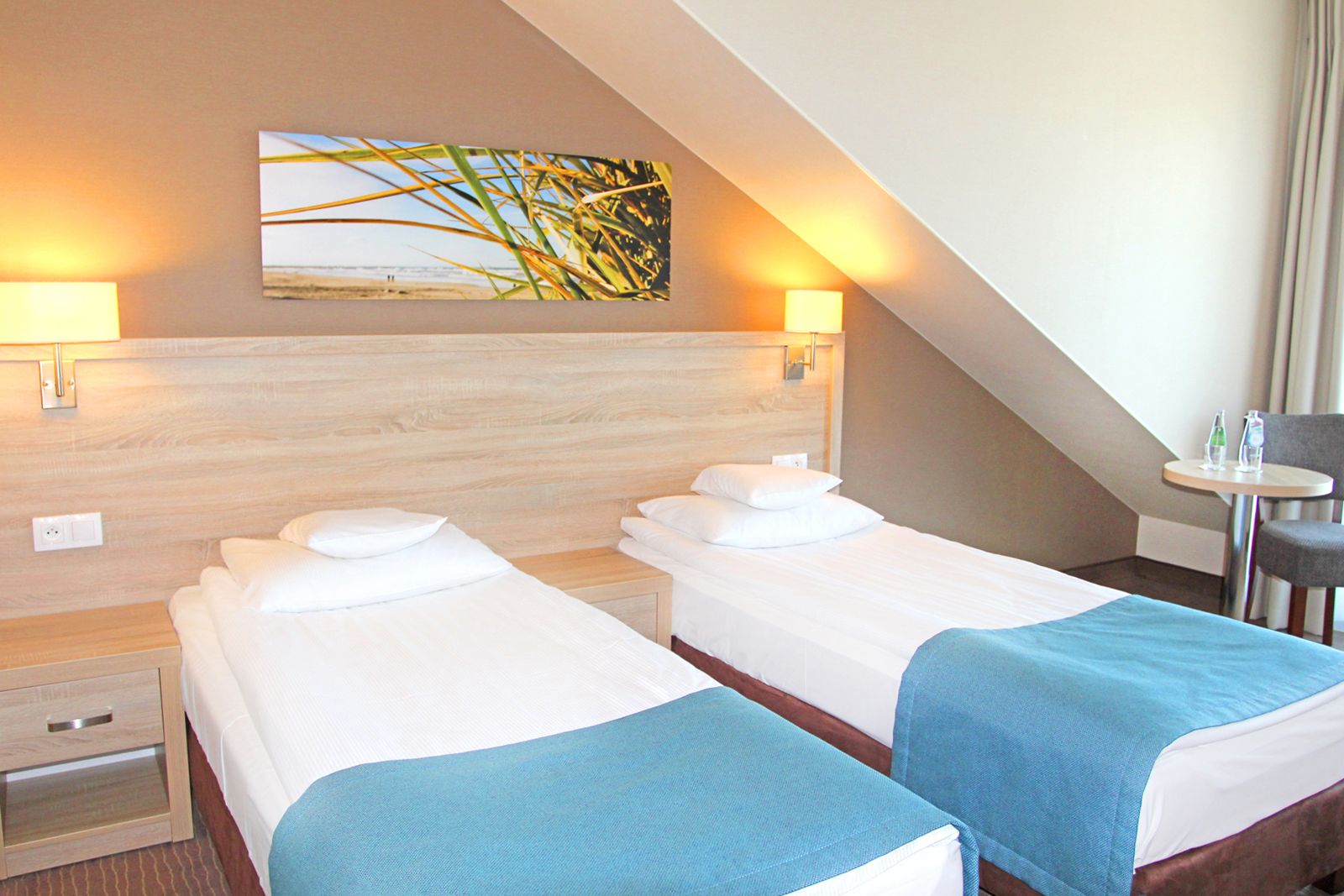A hotel room with two beds, a small table, and a chair. There are wall-mounted lamps and a painting above the beds.
