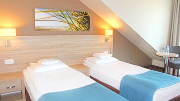 A hotel room with two beds, a small table, and a chair. There are wall-mounted lamps and a painting above the beds.
