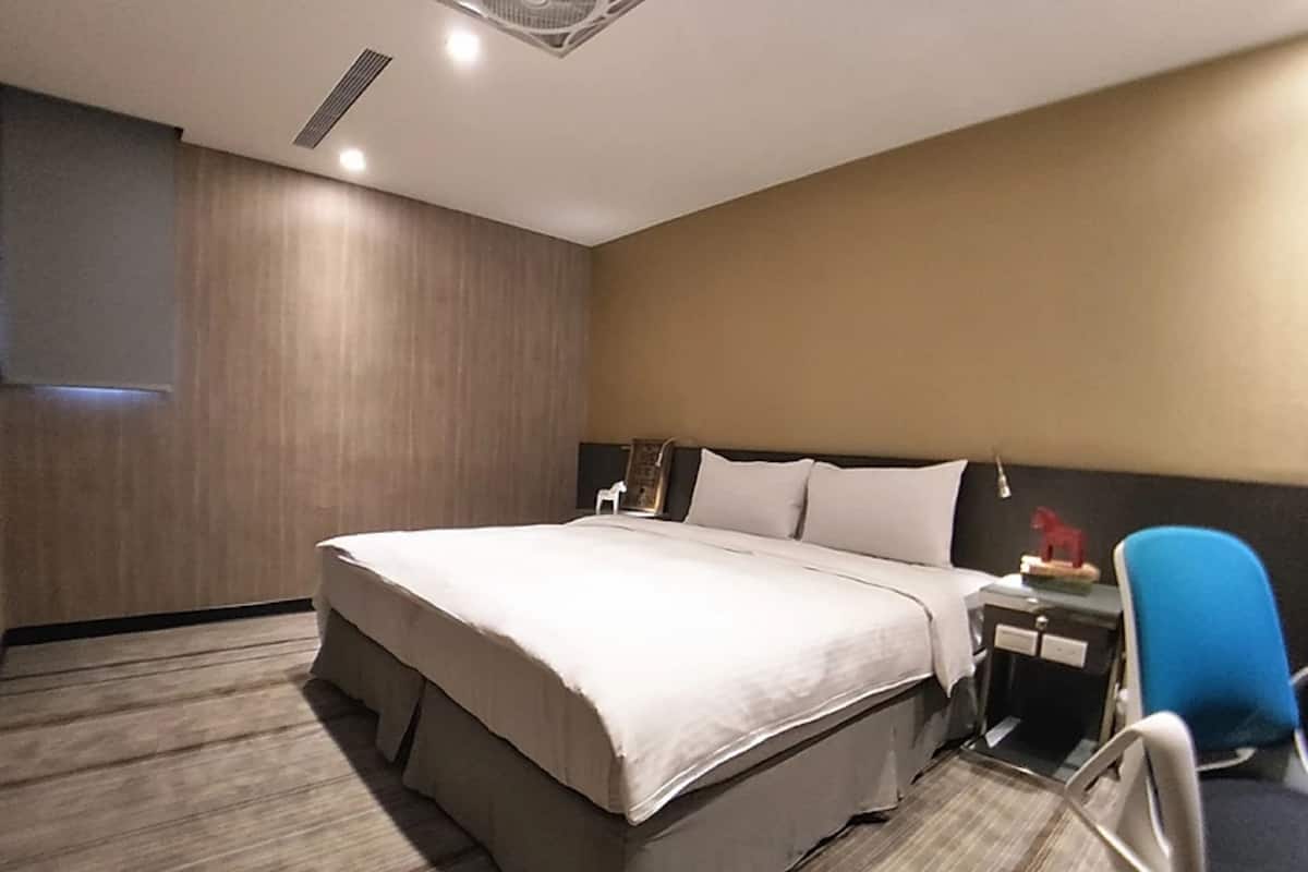 A hotel room with a large bed, a desk with a chair, and a small table with a lamp.