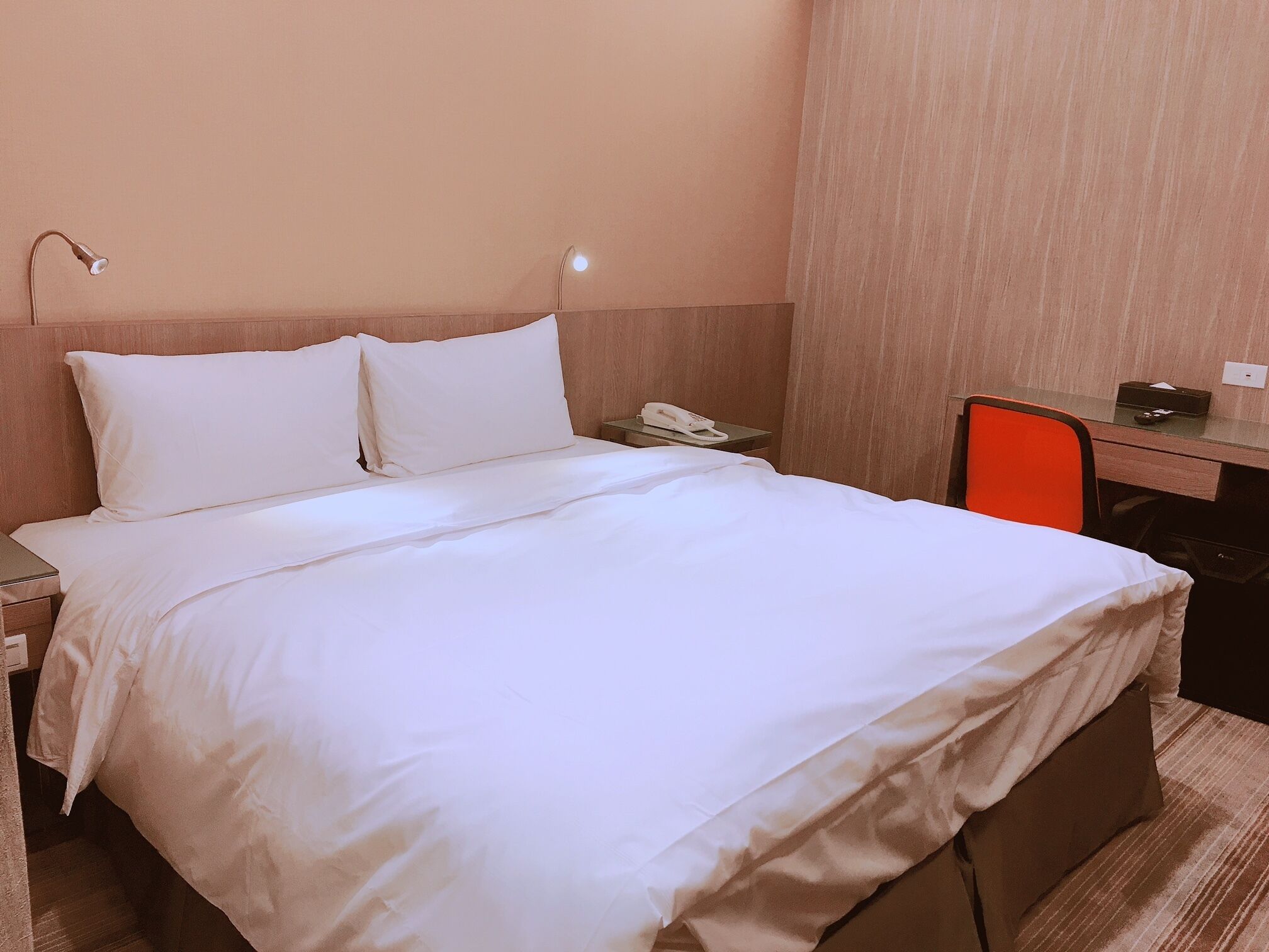 A neatly made bed with white linens, a bedside table with a phone, and a desk with a chair.