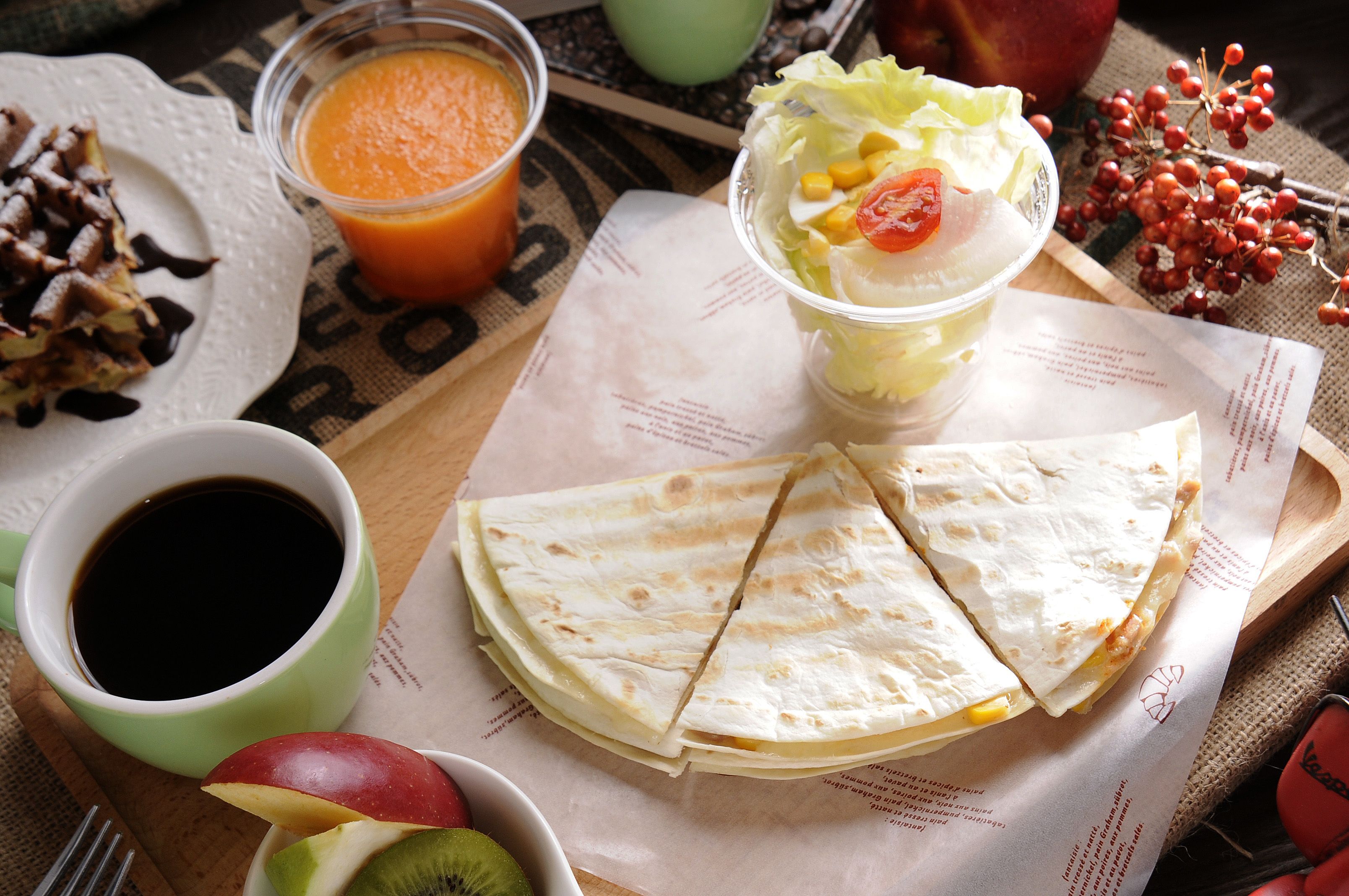 Un plato con quesadillas, una taza de café, un tazón de fruta y un vaso de jugo de naranja sobre una mesa.
