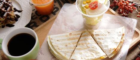 Un plato con quesadillas, una taza de café, un tazón de fruta y un vaso de jugo de naranja sobre una mesa.