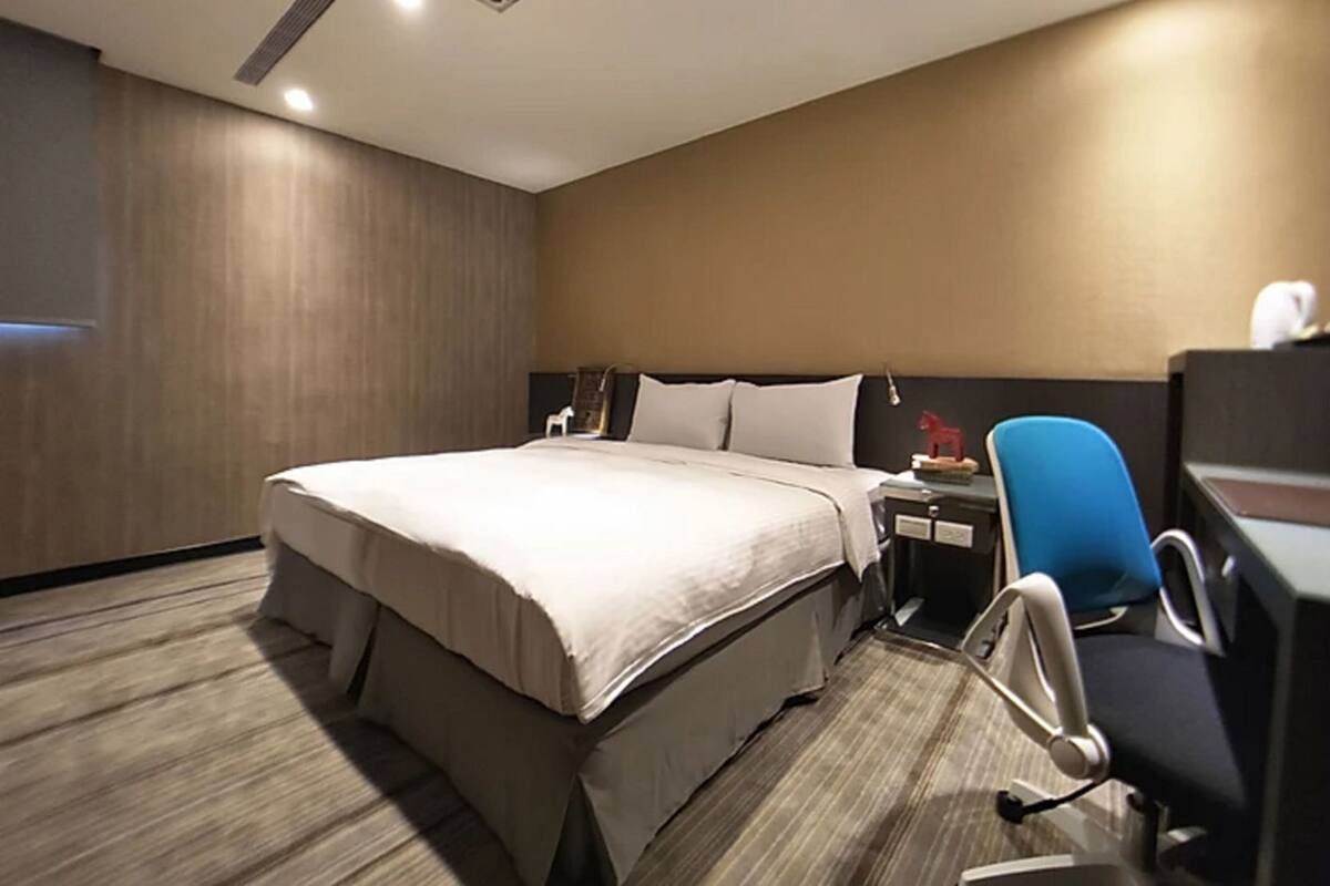 A hotel room with a large bed, a desk with a chair, a nightstand, and a wall-mounted fan.