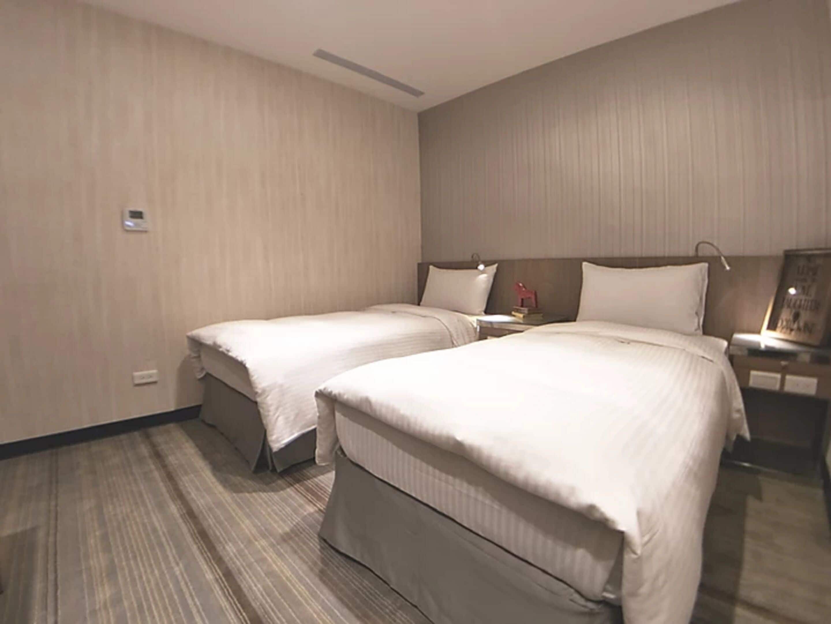 Une chambre d’hôtel avec deux lits simples, une table de chevet avec une lampe et une petite table de nuit avec un livre.