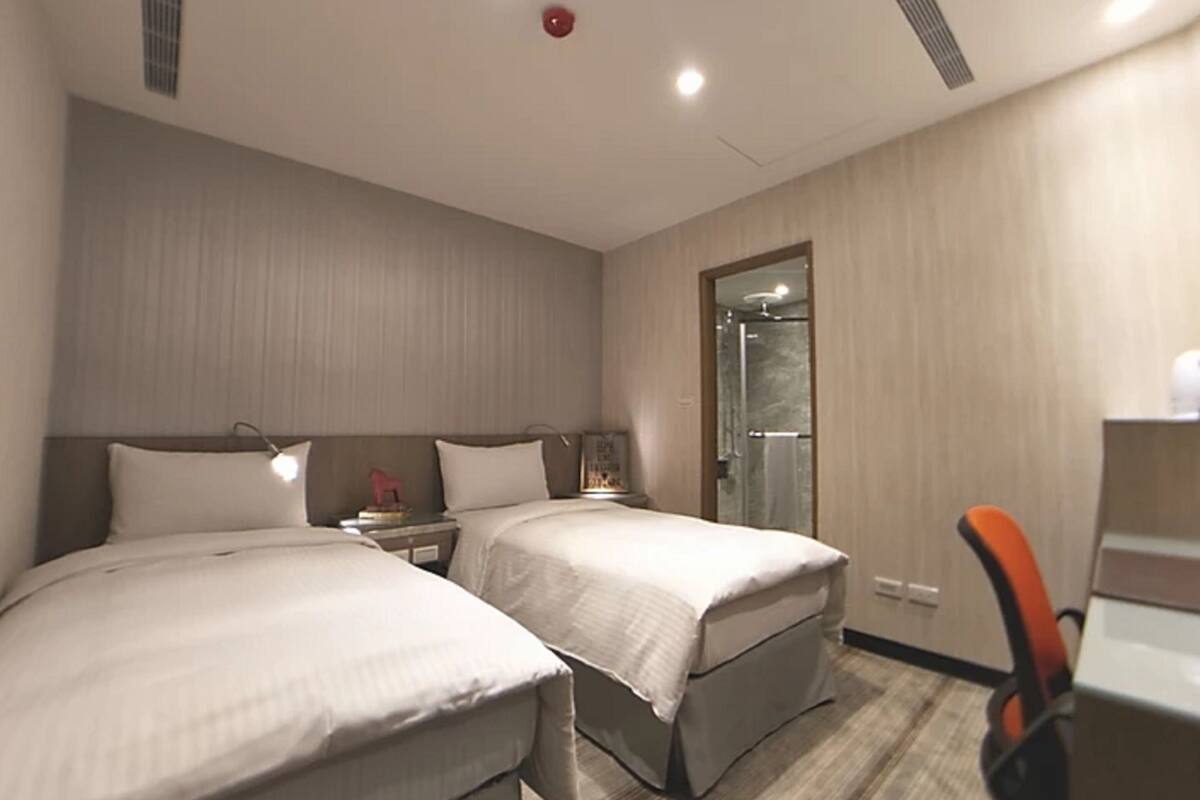 A hotel room with two beds, a desk, and a shower.