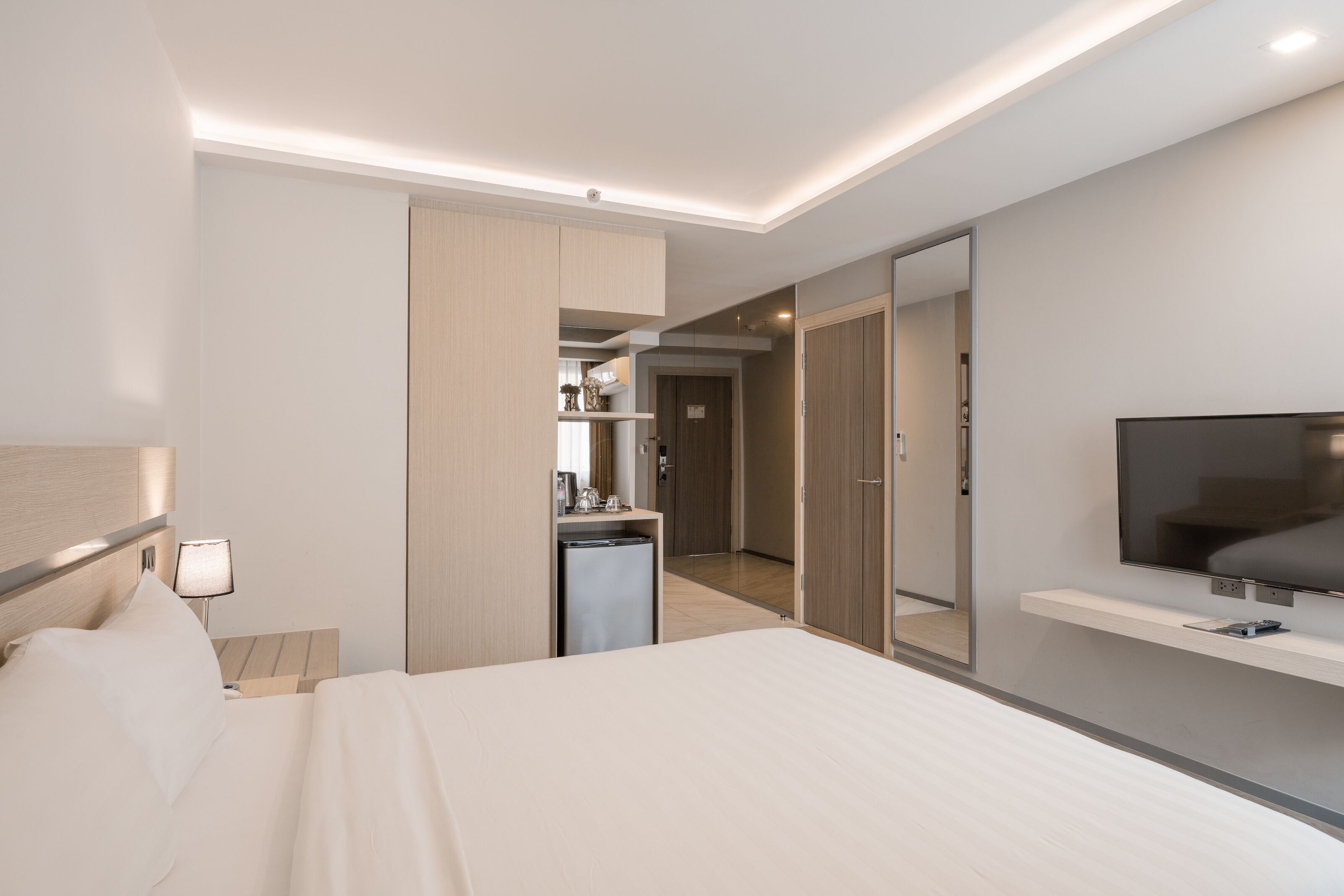 A modern hotel room with a bed, bedside table, lamp, TV, and a small kitchenette area.