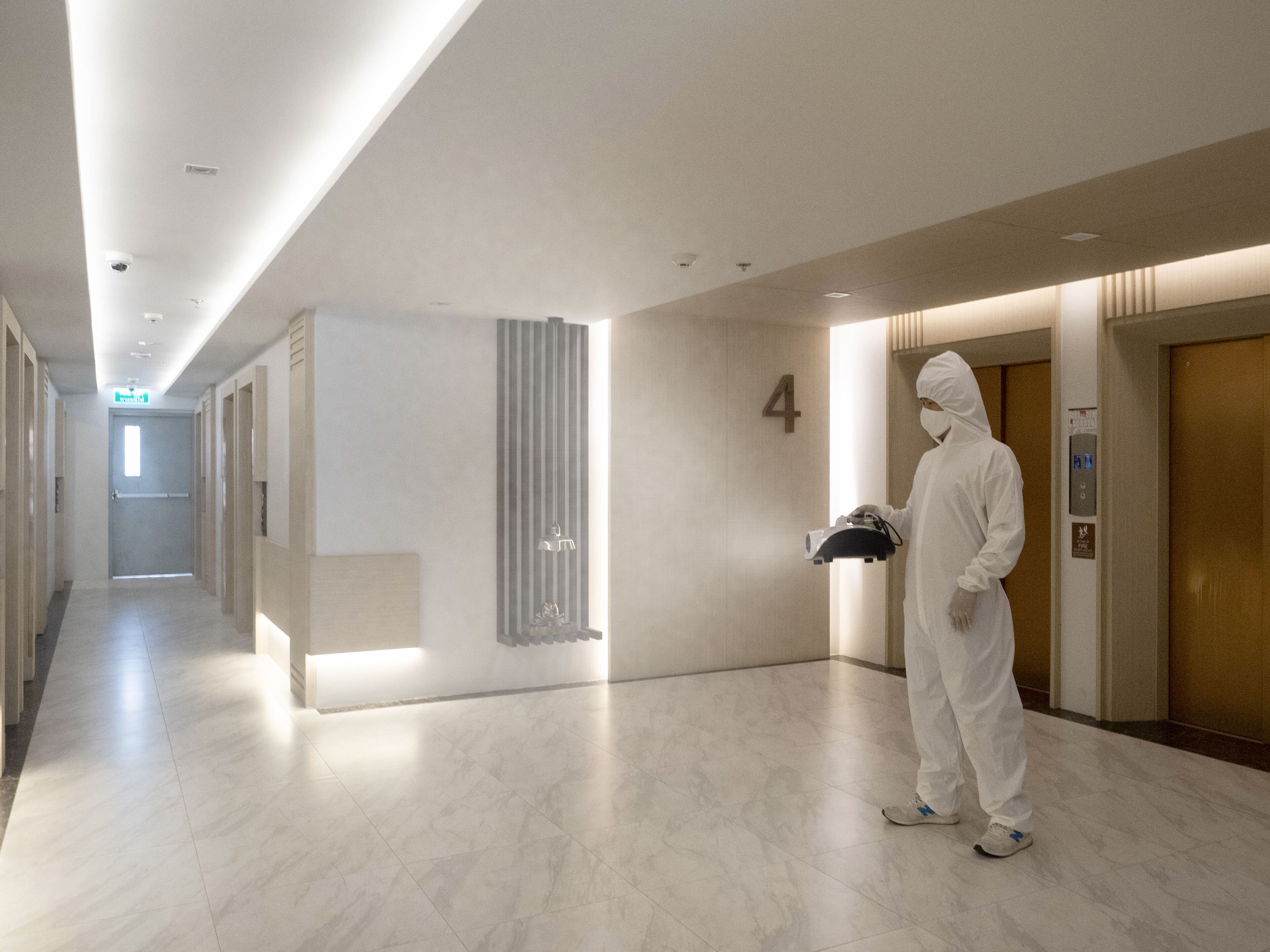 A modern hotel corridor with a clean, minimalist design, featuring a tiled floor, a number '4' sign, and a person in a protective suit carrying a device.