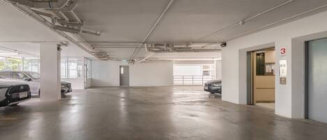 A spacious parking garage with concrete floors and white walls, featuring several parked cars and visible ceiling pipes.