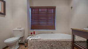 Ein Badezimmer mit Toilette, einer Badewanne vor einer Steinwand, einem Fenster mit Jalousien und einem Waschbecken mit Holzauskleidung.
