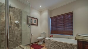 Ein Badezimmer mit Duschkabine aus Glas, Toilette, Badewanne und einem Fenster mit Jalousien.