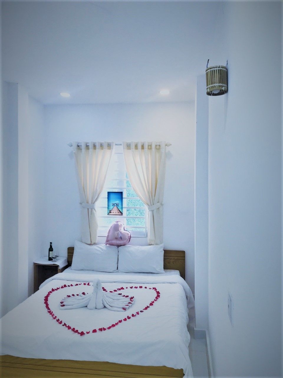 A hotel room with a bed, a nightstand, a window with curtains, and a wall-mounted light fixture.