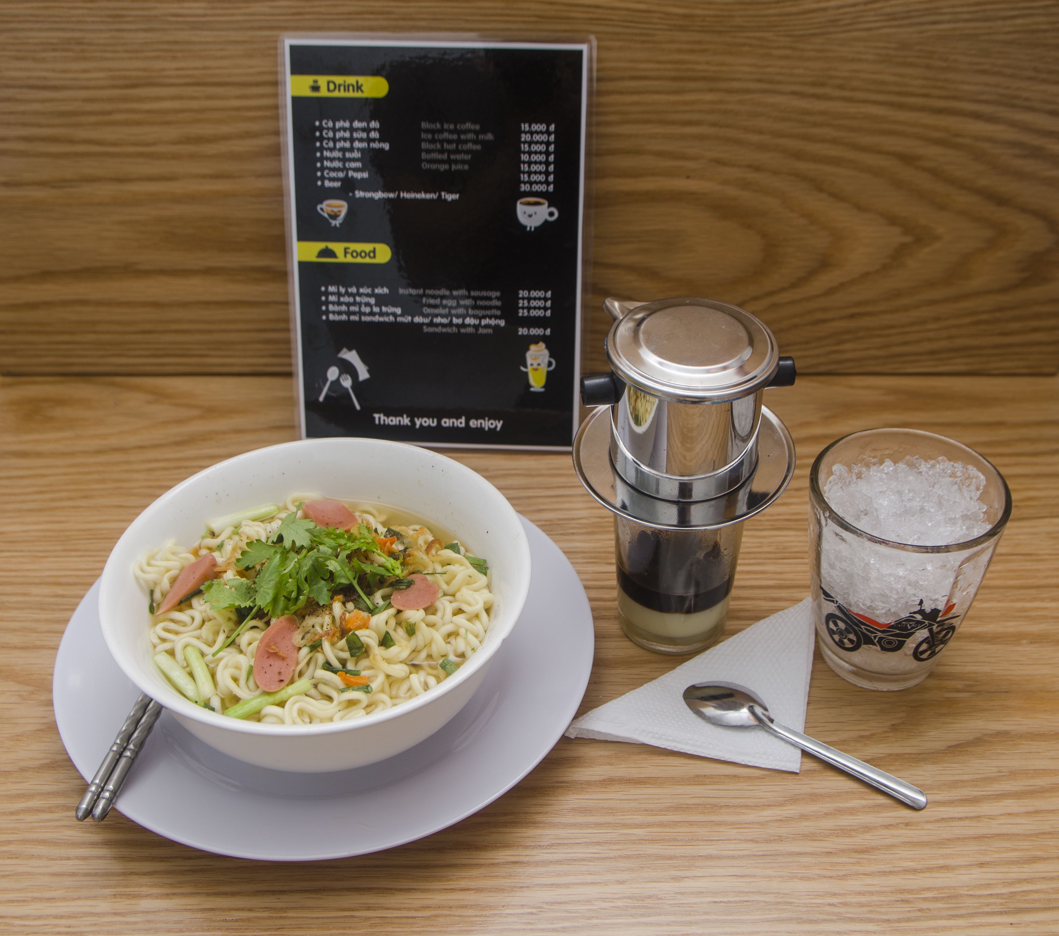 A bowl of ramen noodles with vegetables and meat, a menu listing drinks and food items, a coffee maker, and a glass of iced tea on a wooden table.
