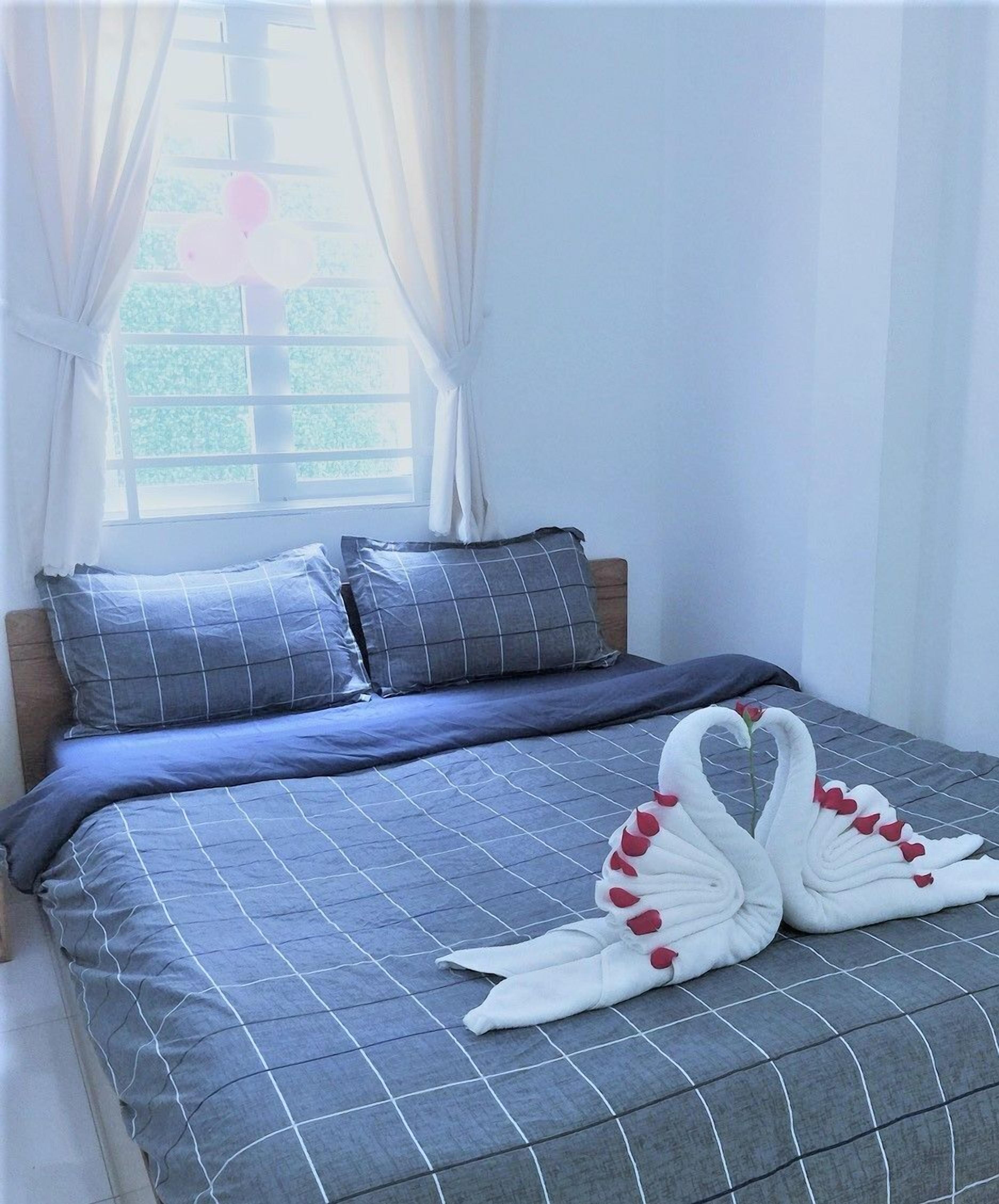 Un lit avec une couverture à carreaux bleus et des oreillers, une fenêtre avec des rideaux et une serviette en forme de cygne décorative posée sur le lit.
