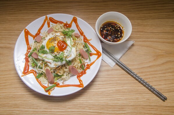 A plate of noodles with a fried egg, greens, and sausage, accompanied by a small bowl of sauce and chopsticks on a wooden table. - H Best Hostel Cafe Saigon (Ho Chi Minh City)