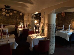 A dimly lit restaurant with stone walls and mosaic tile accents. Tables are set with candles and white tablecloths.