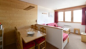 A compact room with a wooden dining table, chairs, a small kitchenette, and a window with curtains.