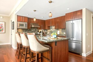 A modern kitchen with wooden cabinets, a stainless steel refrigerator, a marble island with bar stools, and pendant lighting.