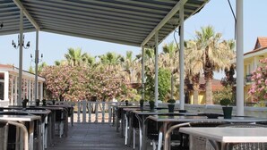An outdoor cafe with tables and chairs under a covered area, surrounded by palm trees and flowering plants. - Hierapolis Termal Hotel (Denizli)