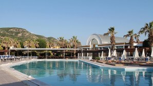 A swimming pool with lounge chairs and umbrellas, surrounded by palm trees and a building with a dome roof. - Hierapolis Termal Hotel (Denizli)
