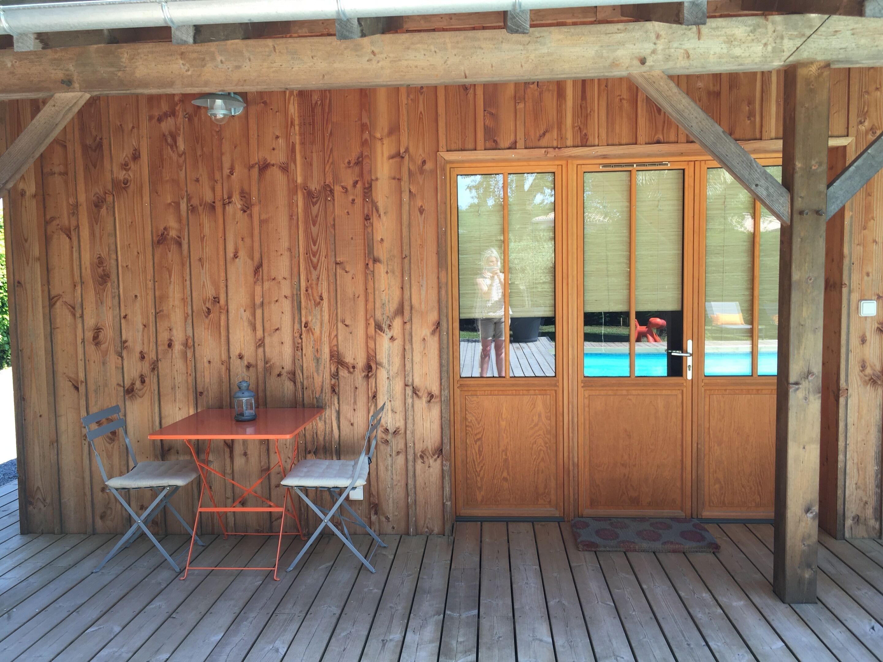 A wooden deck with a table and two chairs, a covered area with a door, and a visible swimming pool in the background.