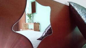 Un espejo de forma original refleja una habitación con un aparador de madera, una planta y un ventanal.