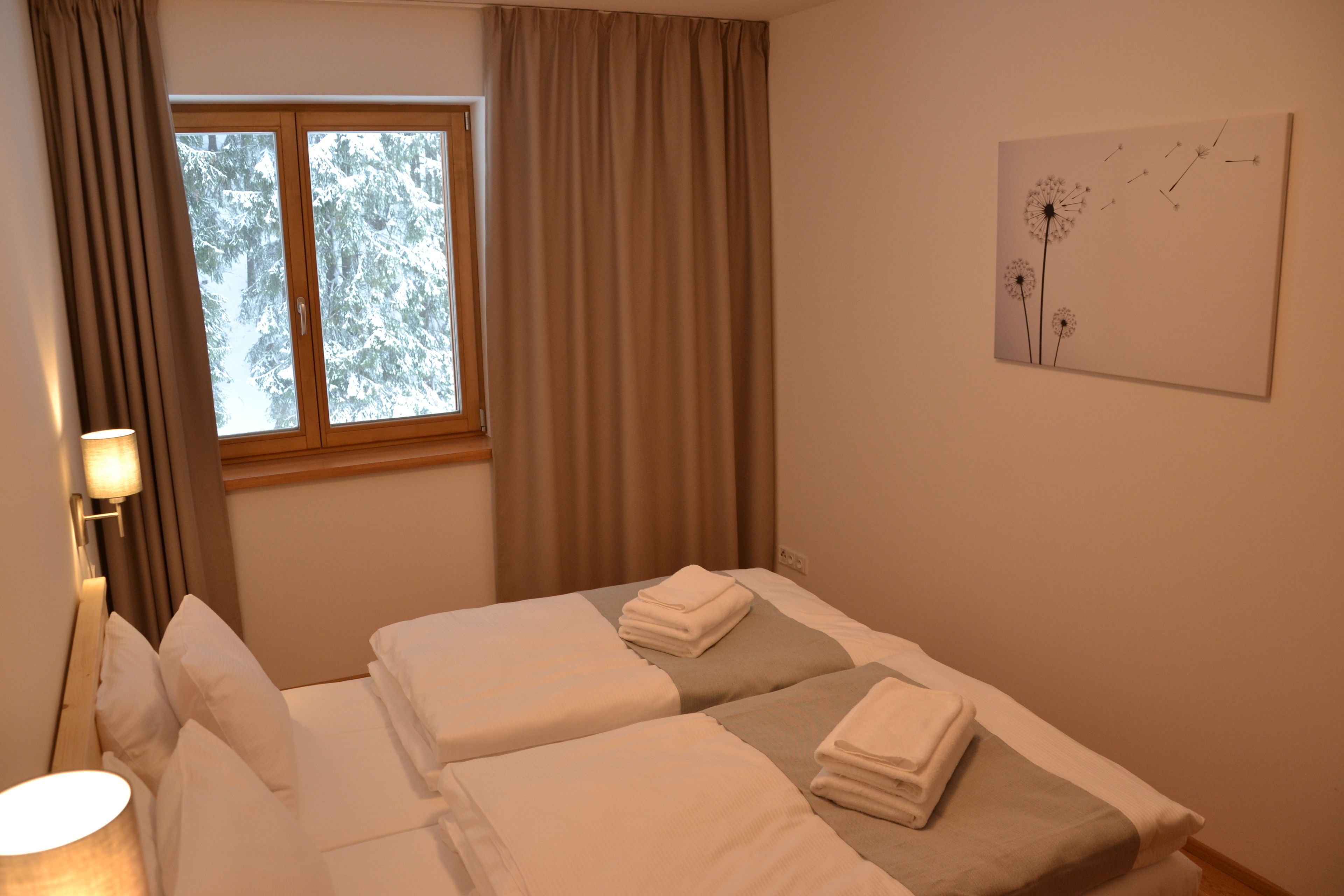 A hotel room with two beds, a window with curtains, a wall-mounted lamp, and a painting of dandelions.
