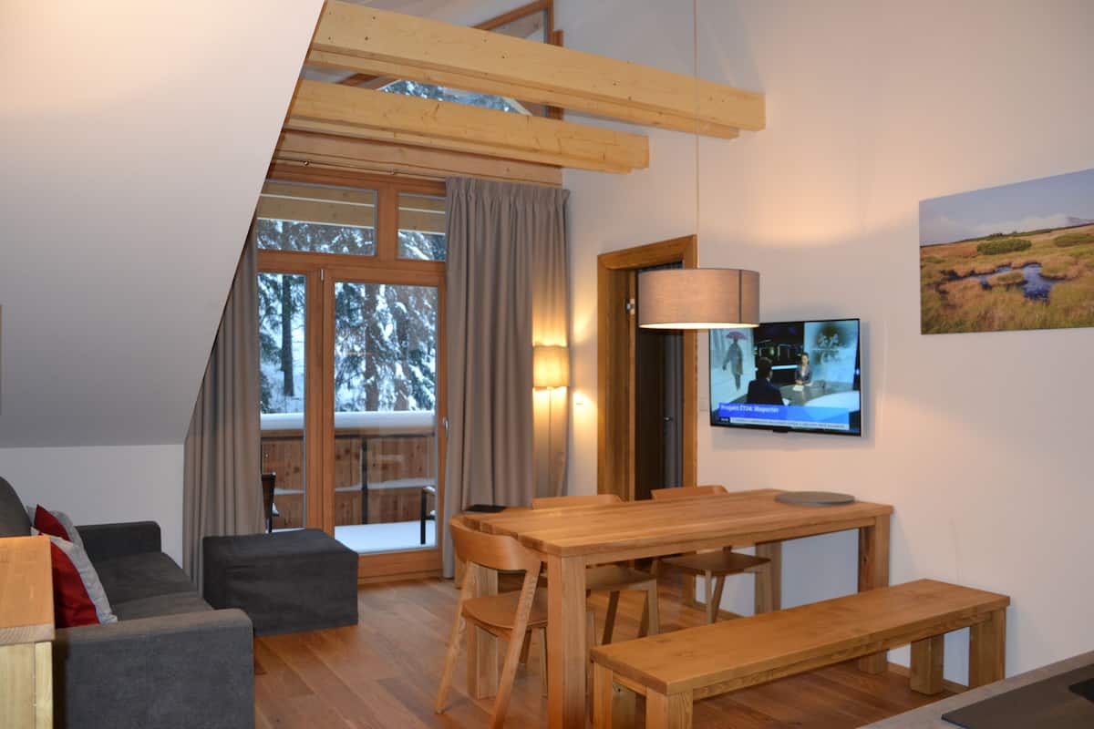 A cozy living room with a wooden dining table, a sofa, and a TV mounted on the wall.
