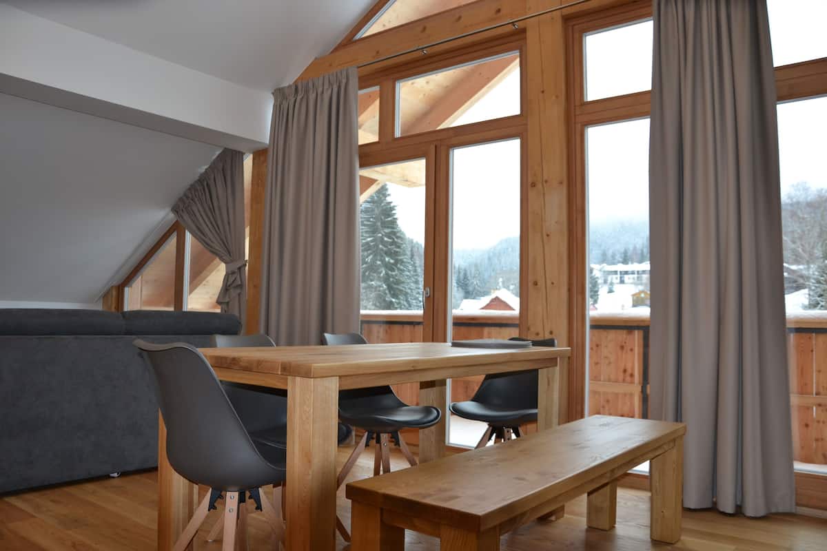 A cozy room with a wooden dining table, black chairs, a grey sofa, and large windows offering a view of snowy mountains.