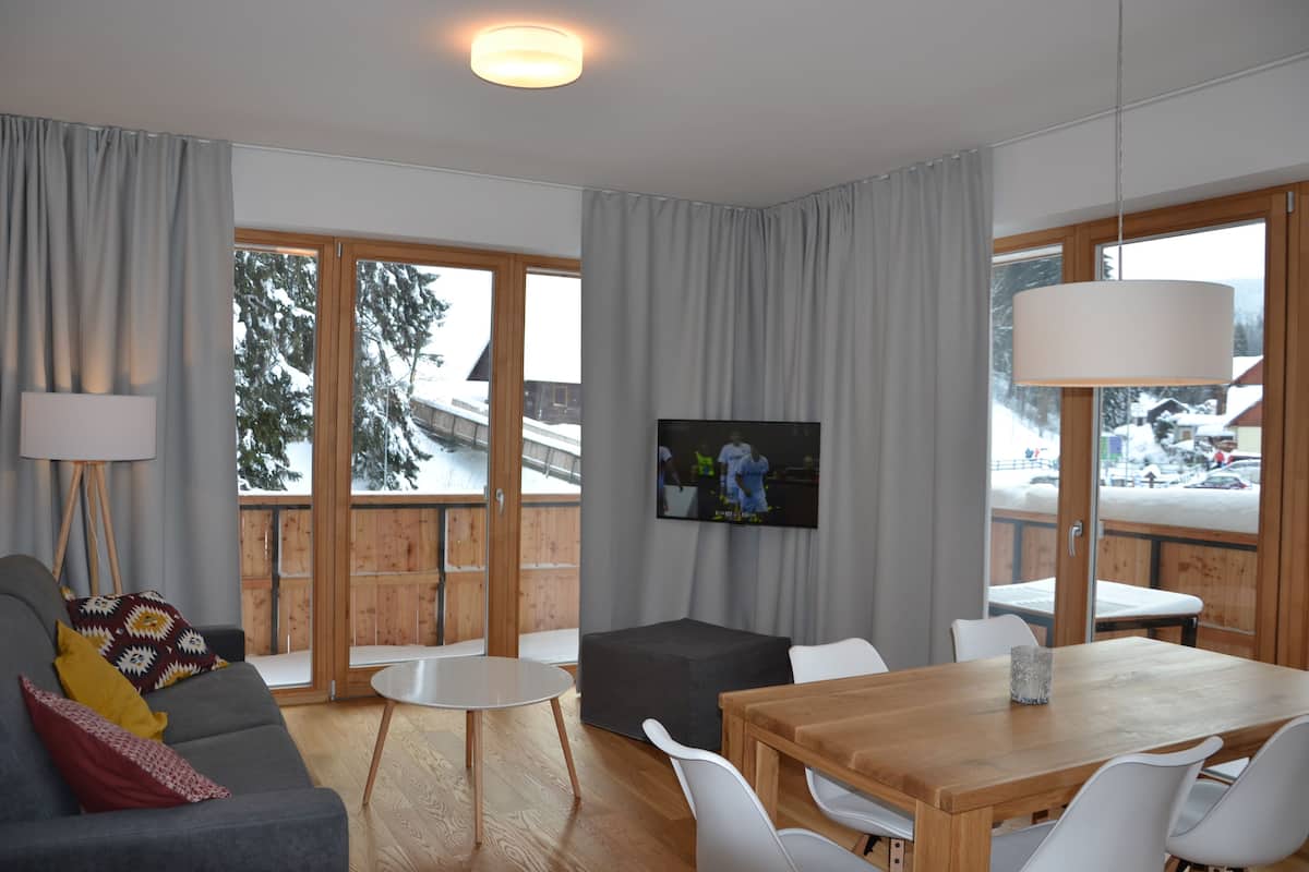 A modern living room with a sofa, coffee table, and dining table. There is a TV mounted on the wall and a large window with a view of a snowy landscape.