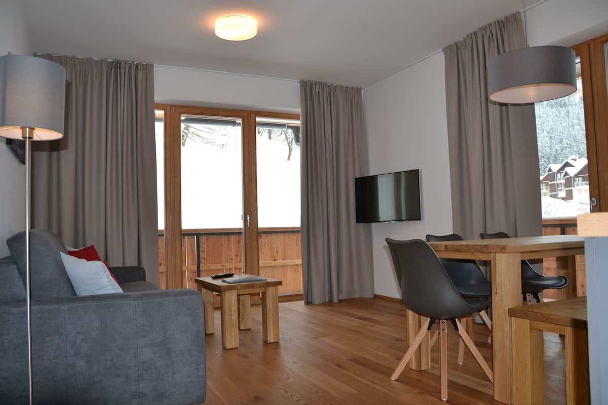 A modern living room with a sofa, a dining table, and chairs. There is a flat-screen TV mounted on the wall.